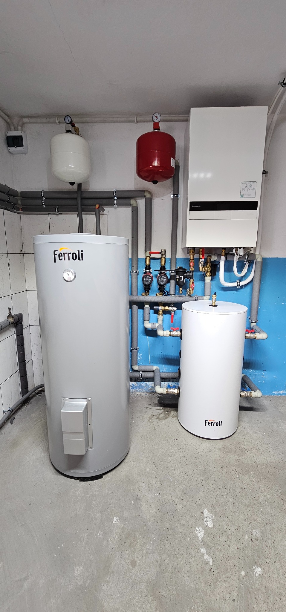 Instalacja grzewcza w kotłowni: szary zasobnik ciepłej wody Ferroli z widocznym manometrem, biały zasobnik ciepłej wody Ferroli, wiszący biały piec grzewczy Panasonic, czerwony zbiornik wyrównawczy...
