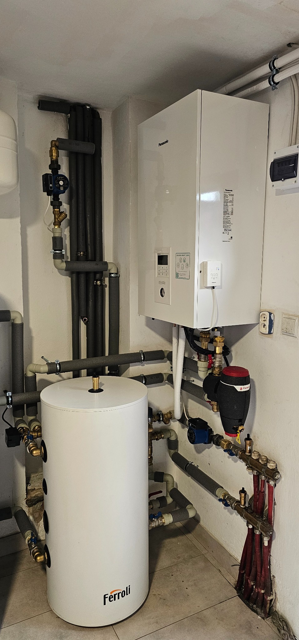 Biały zasobnik ciepłej wody Ferroli i wiszący na ścianie biały moduł Panasonic z licznymi rurami, zaworami i pompami w kotłowni.
