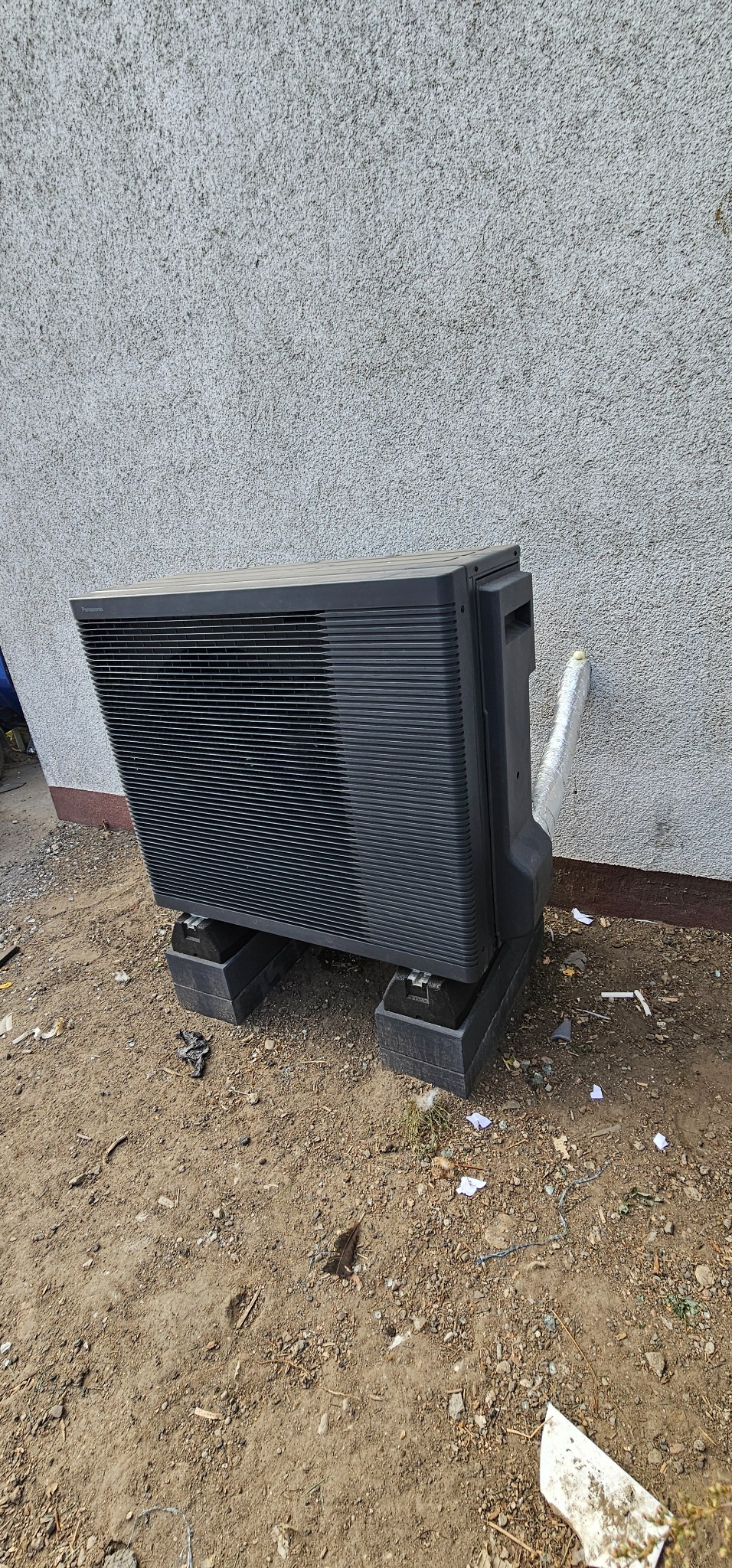 Zewnętrzna jednostka klimatyzacyjna marki Panasonic zamontowana na betonowych podstawach przy szarej ścianie budynku, z widoczną izolacją rury.