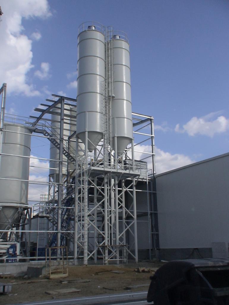 Silosy przemysłowe z blachy falistej, połączone stalową konstrukcją wsporczą i schodami, na tle błękitnego nieba z obłokami.