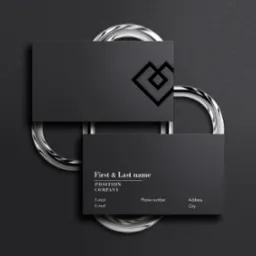 Dwie czarne wizytówki ułożone jedna na drugiej, podparte przez dwa srebrne pierścienie na ciemnoszarym teksturowanym tle. Górna wizytówka ma wycięte geometryczne logo.