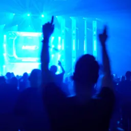 Tłum ludzi z podniesionymi rękami na koncercie oświetlonym intensywnym niebieskim światłem, widoczna scena z ekranem wyświetlającym logo.