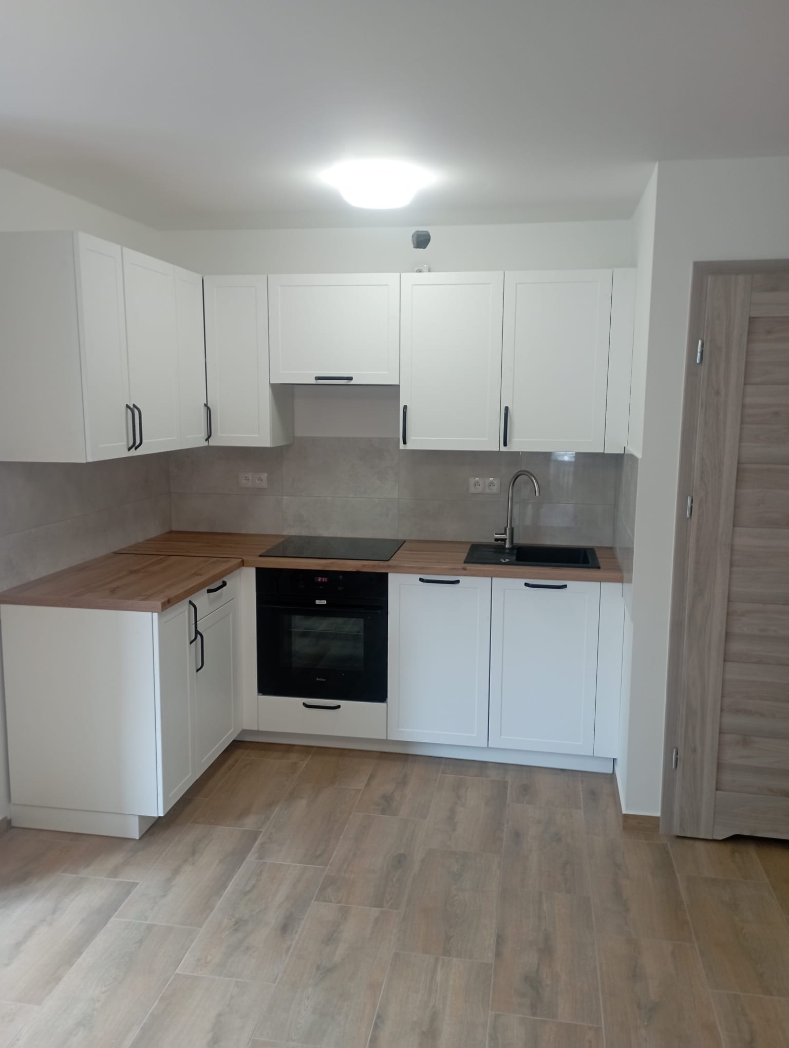 Jasna kuchnia z białymi szafkami i drewnianym blatem, czarny zlew i płyta indukcyjna, płytki ścienne w odcieniach szarości, podłoga wyłożona panelami imitującymi drewno.