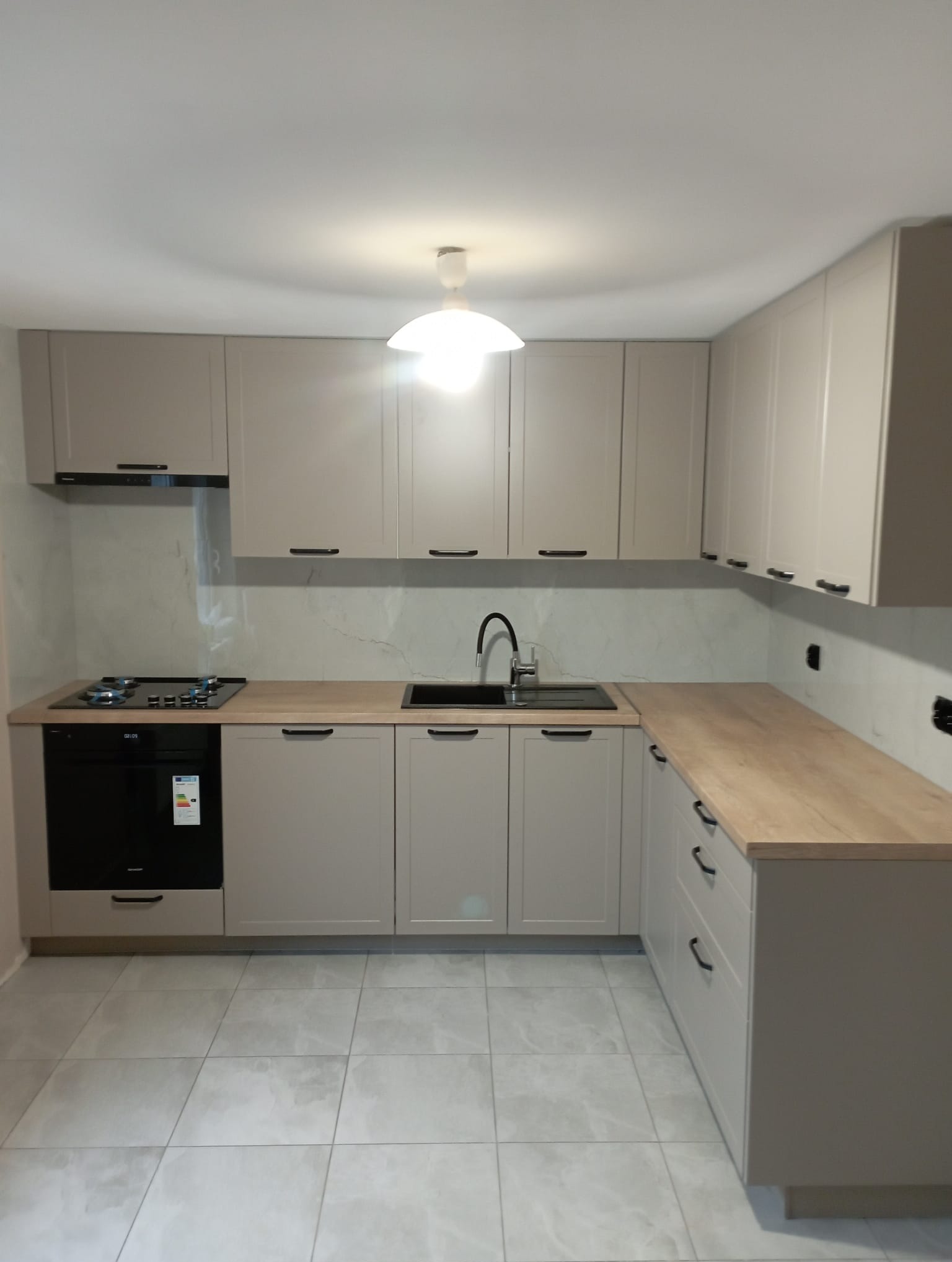 Jasna kuchnia na wymiar z drewnianym blatem, czarnym zlewem i płytą gazową, uchwyty w kolorze czarnym, szare płytki podłogowe.