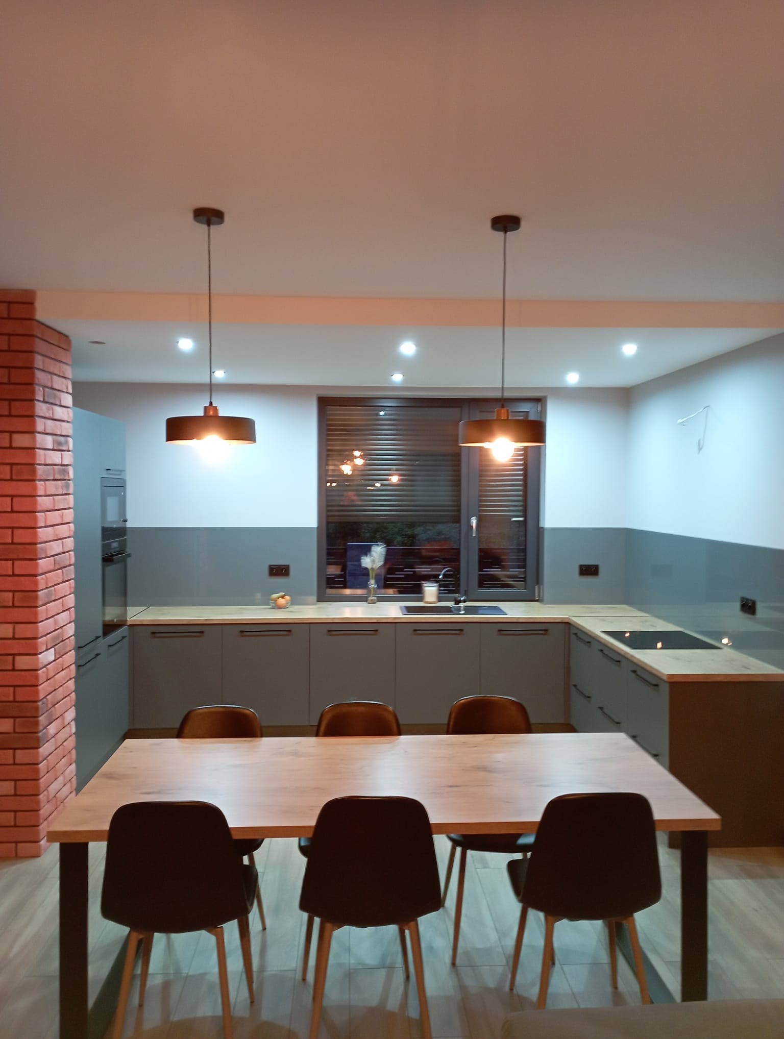 Nowoczesna kuchnia w odcieniach szarości z drewnianym blatem, oświetlona wiszącymi lampami nad stołem jadalnym z czarnymi krzesłami; ściana z cegły po lewej stronie.
