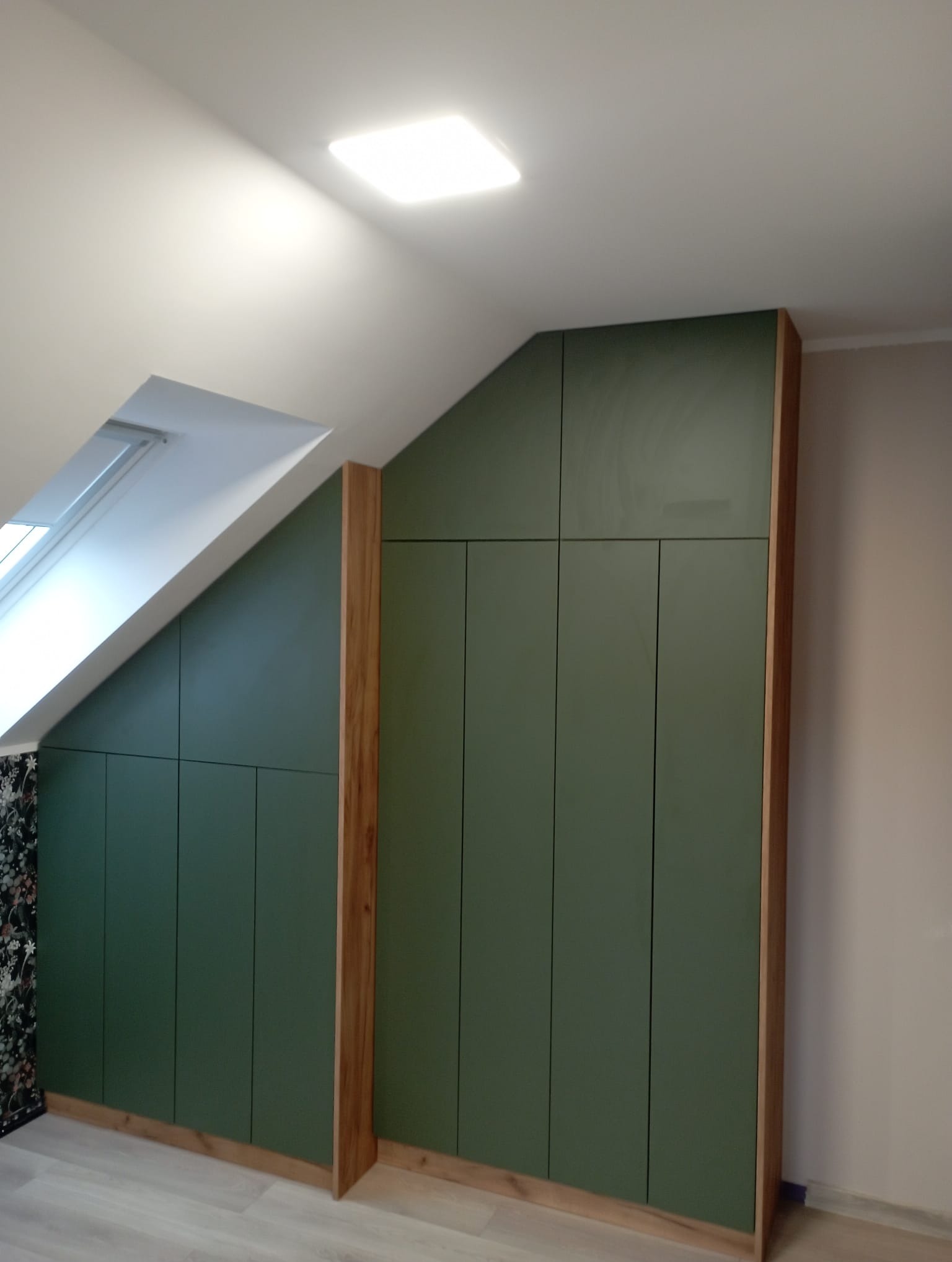 Zielona szafa wnękowa z drewnianymi elementami dopasowana do skosu poddasza, widoczne okno dachowe i wzorzysta tapeta