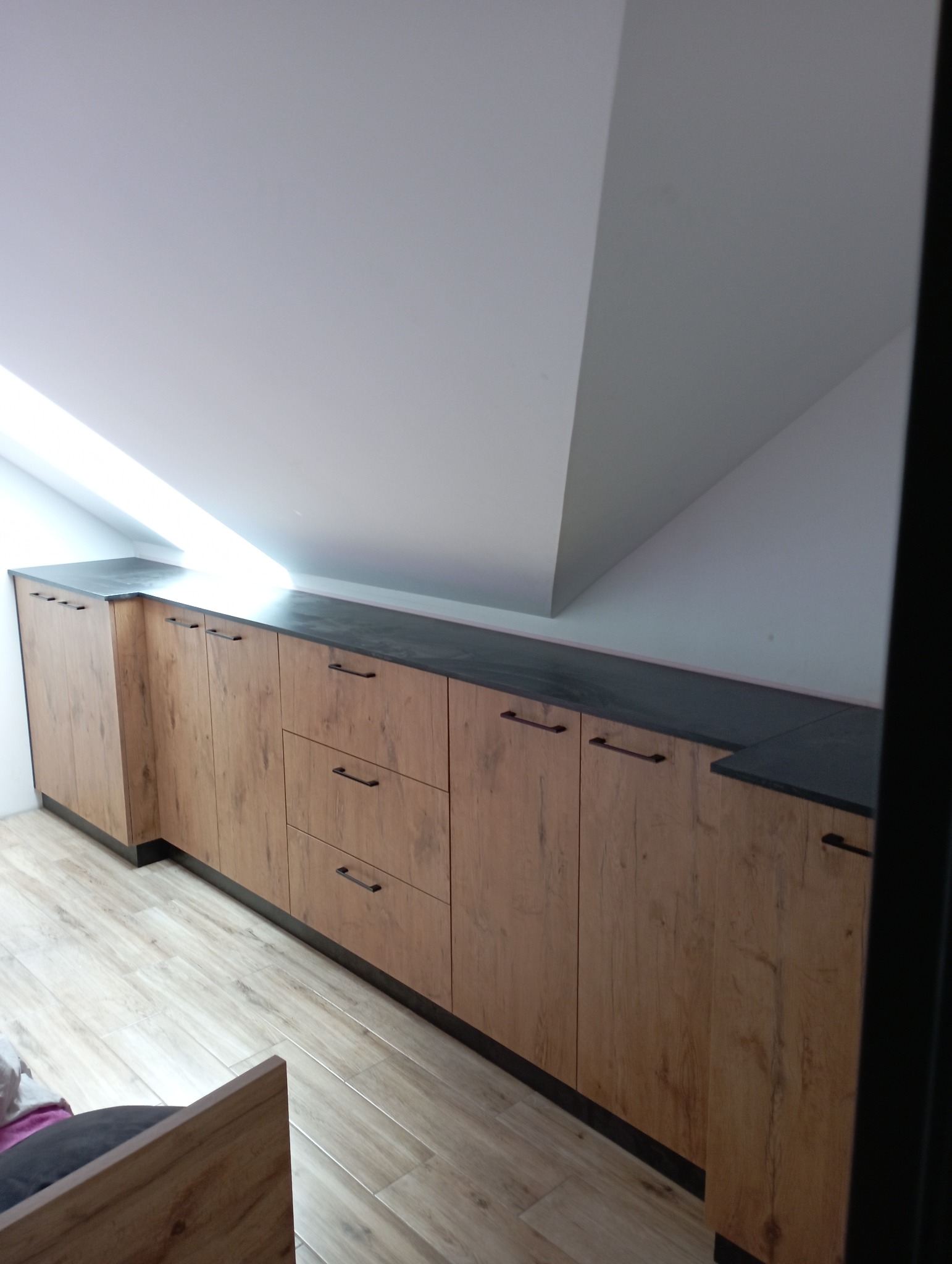 Zabudowa wnęki na poddaszu z drewnopodobnymi frontami i ciemnym blatem, widoczne poziome uchwyty, jasna podłoga imitująca drewno.