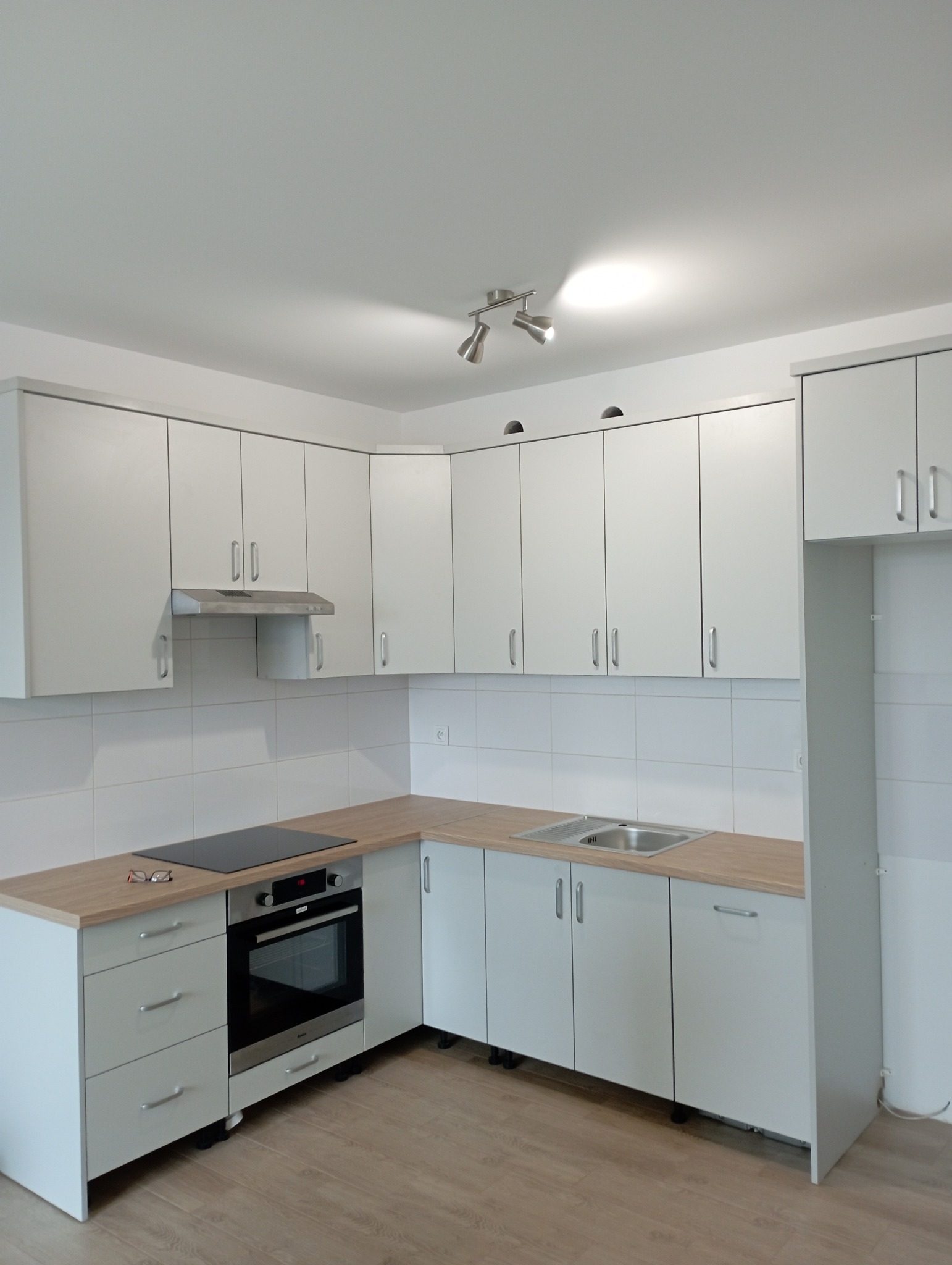 Jasna, narożna zabudowa kuchenna z drewnianym blatem, płytą indukcyjną i piekarnikiem, uzupełniona o białe płytki na ścianie, oświetlenie sufitowe i okulary leżące na blacie.