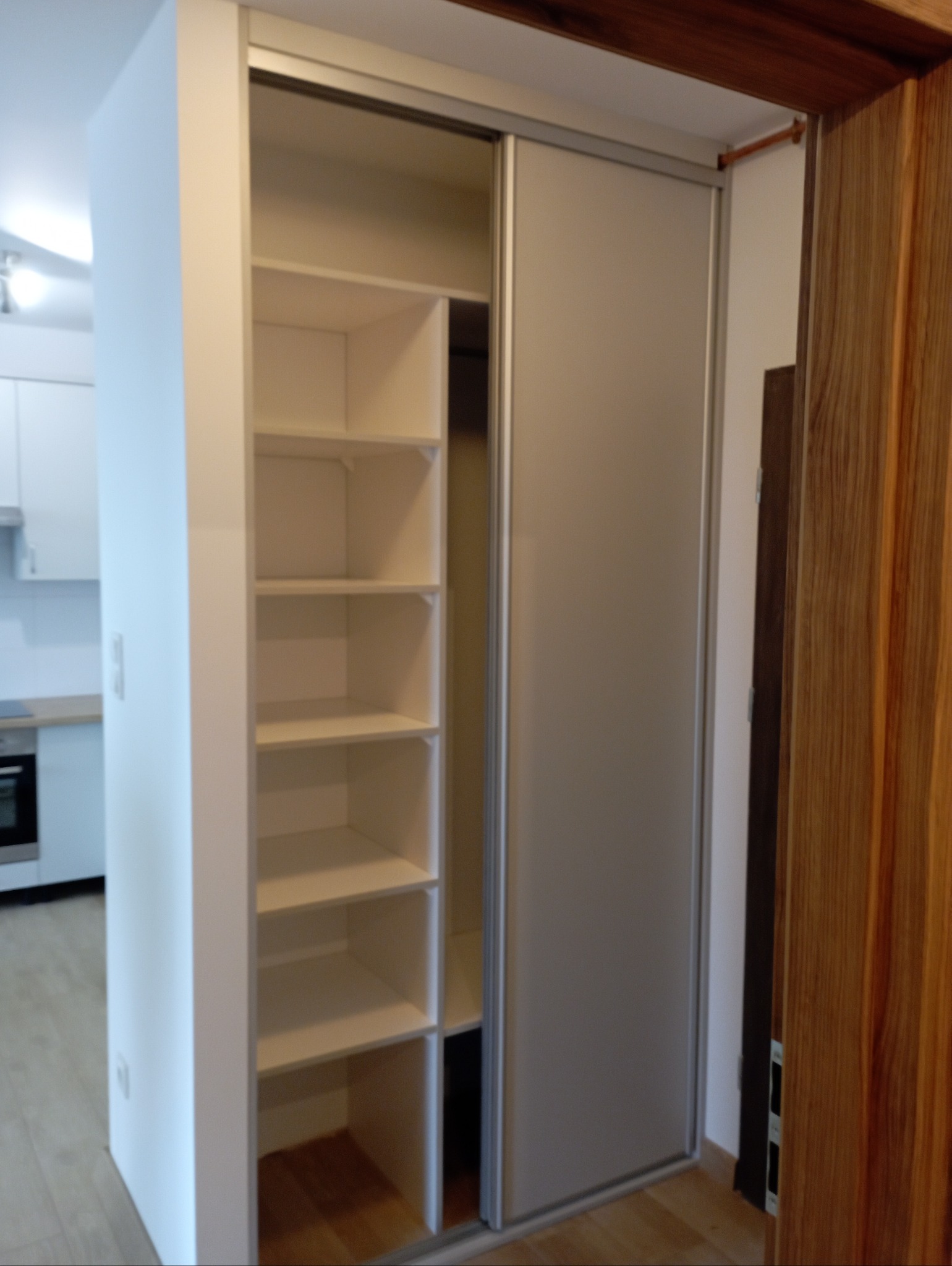 Wnętrze szafy wnękowej z przesuwnymi drzwiami, widoczne półki i fragment kuchni w tle.