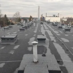 Widok z góry na płaski dach pokryty papą z licznymi kominkami wentylacyjnymi i instalacją odgromową, na tle zabudowań miejskich w pochmurny dzień.