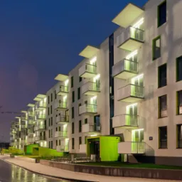 Nowoczesny budynek wielorodzinny z białymi elewacjami i zielonymi akcentami, widoczny po zmroku, z oświetlonymi balkonami i mokrą nawierzchnią ulicy odbijającą światła, w tle żurawie budowlane.