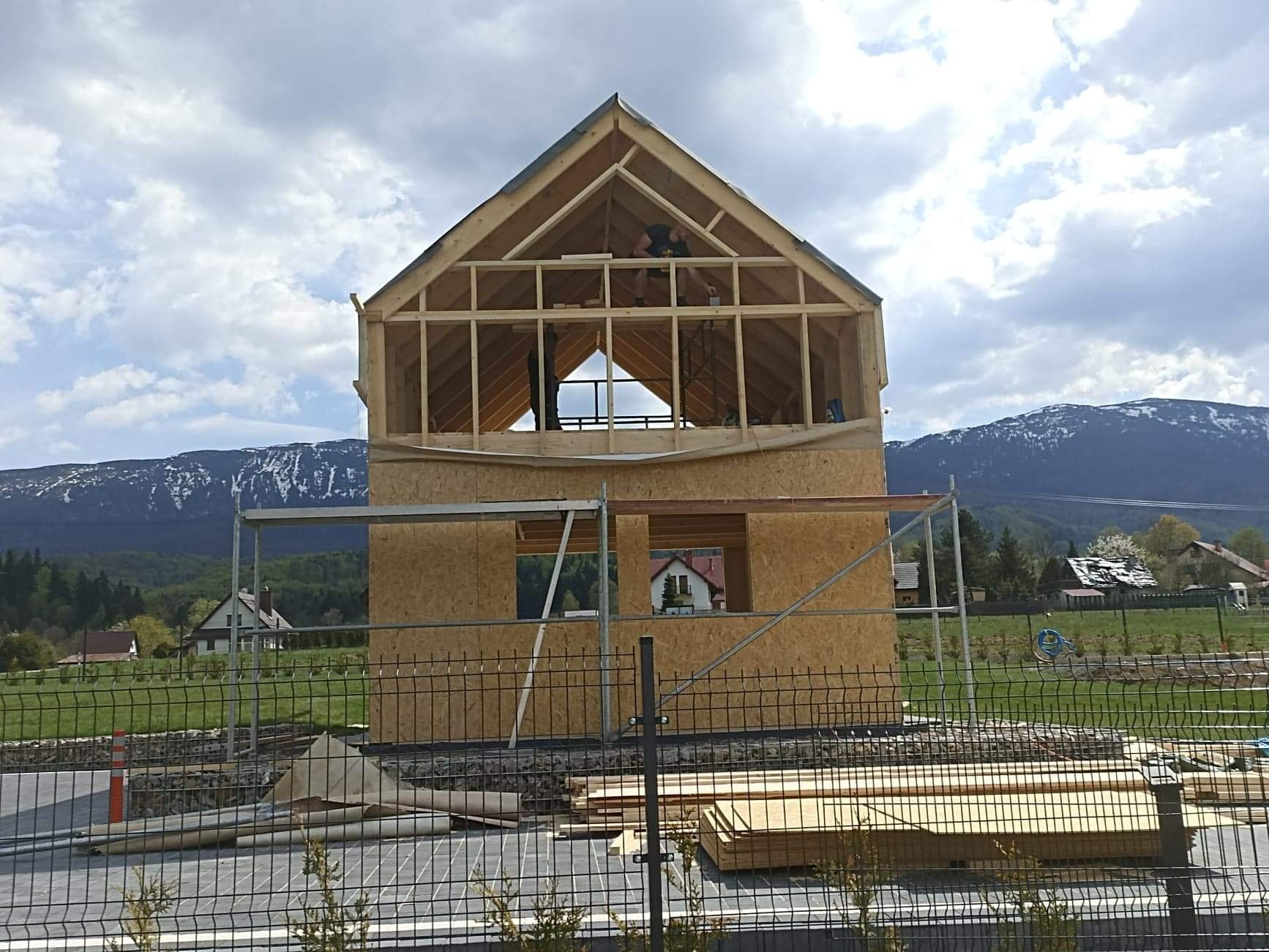Surowy szkielet dwukondygnacyjnego domu w trakcie budowy, widoczne drewniane elementy konstrukcyjne i płyty OSB, w tle góry ze śnieżnymi szczytami pod pochmurnym niebem.
