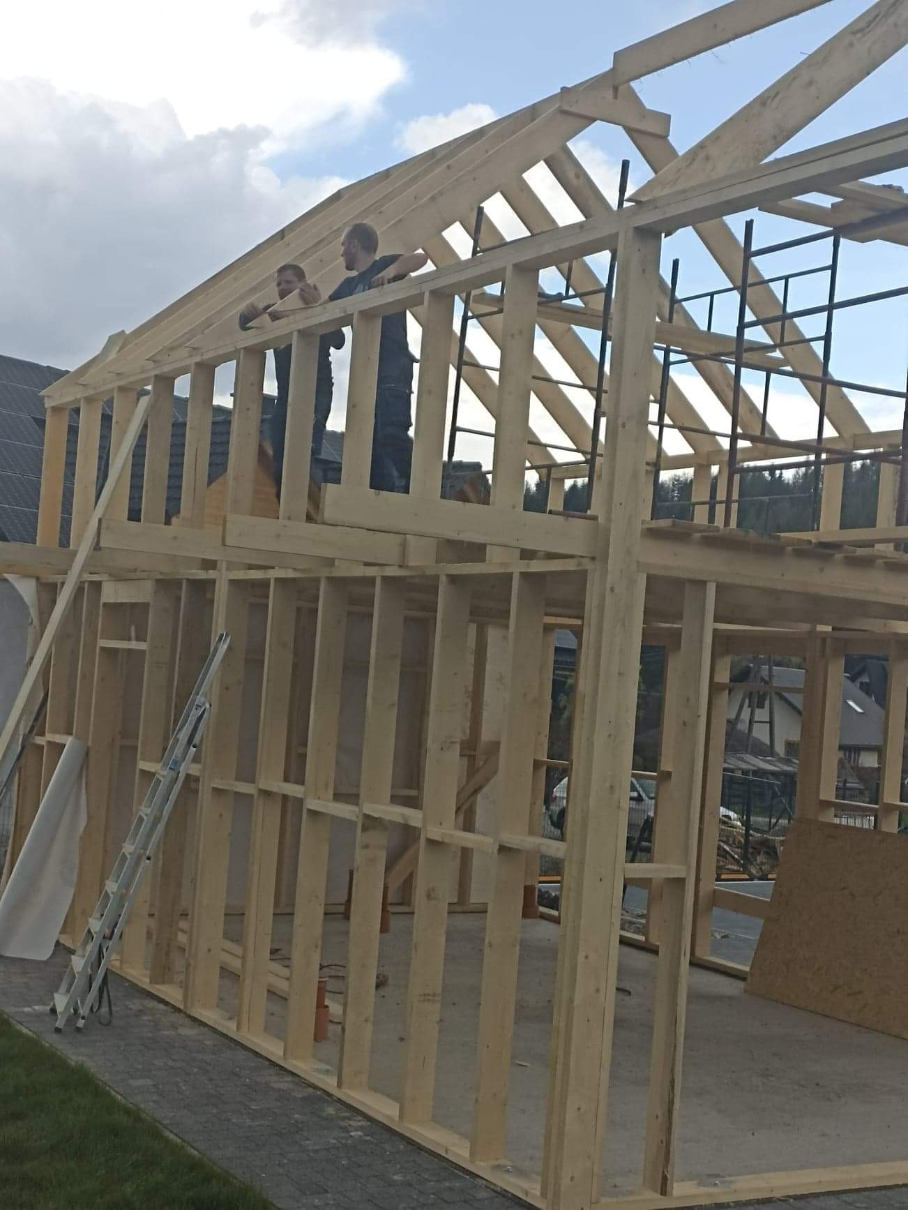 Konstrukcja szkieletowa domu w trakcie budowy, dwóch robotników na górze, widoczne drewniane elementy konstrukcyjne, drabina oparta o ścianę, w tle domy i drzewa.