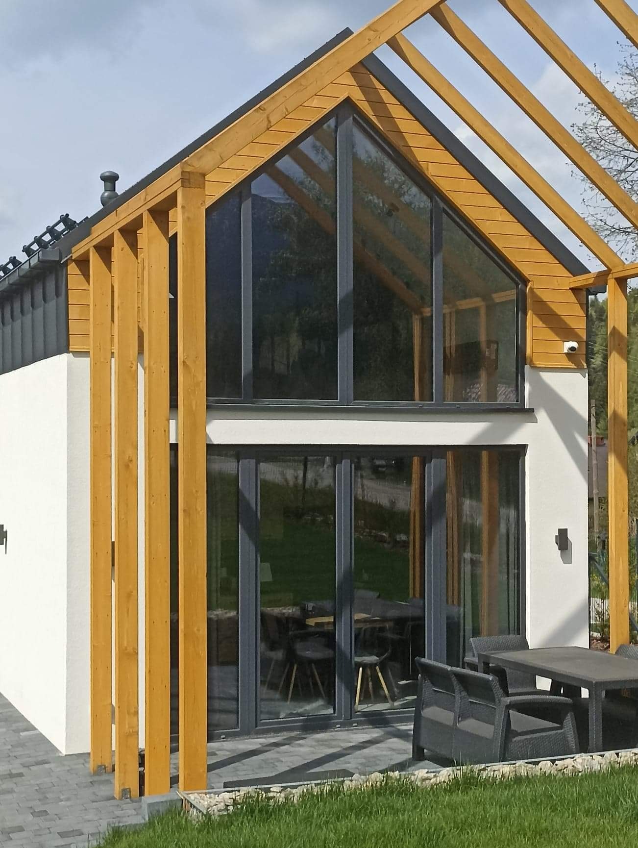 Nowoczesny dom szkieletowy z dużymi przeszkleniami i drewnianą konstrukcją, widok z zewnątrz na taras z meblami ogrodowymi.