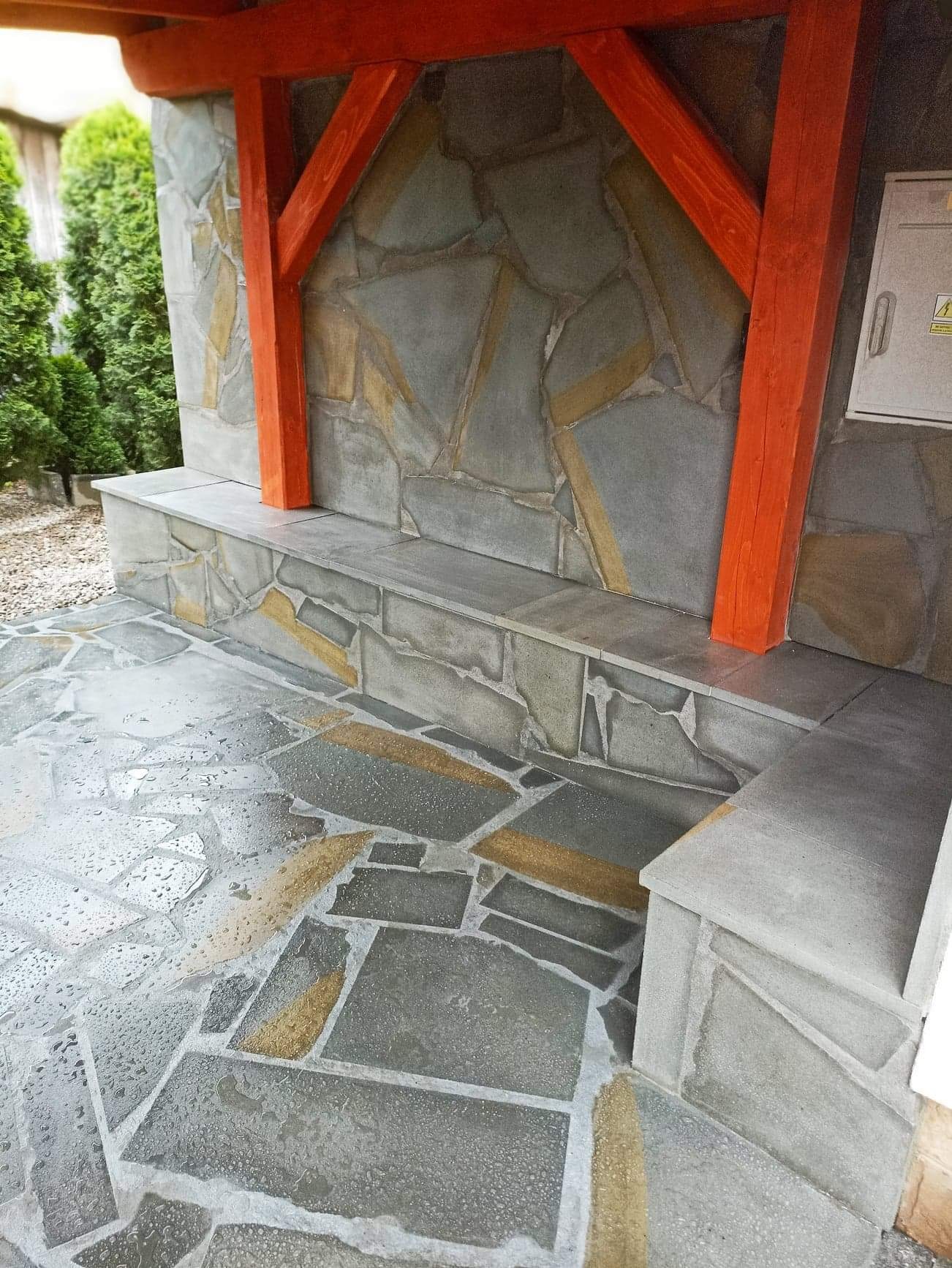Kamienna elewacja z elementami dekoracyjnymi w odcieniach szarości i beżu, z pomarańczowymi drewnianymi belkami podtrzymującymi zadaszenie, oraz kamienną ławką i podłogą z nieregularnych płyt.