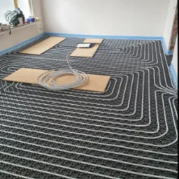 Rozłożone rury ogrzewania podłogowego na czarnej macie izolacyjnej, zabezpieczone folią przy ścianach, z drewnianymi płytami i zwiniętym szarym przewodem na wierzchu, w surowym wnętrzu.