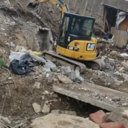 Żółta koparka CAT 305E pracująca na terenie z gruzem i resztkami budynków, widoczny fragment starego muru i sterta śmieci, drzewa w tle.