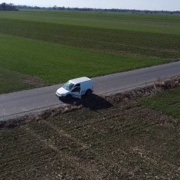 ElektroGriner - Biały van z otwartymi drzwiami stoi na wąskiej drodze asfaltowej, otoczony polami uprawnymi w słoneczny dzień. Zdjęcie z lotu ptaka.