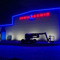 ElektroGriner - Budynek firmy „Induserwis” z neonowym oświetleniem elewacji i podnośnikiem koszowym na tle nocnego nieba, oświetlony reflektorami.