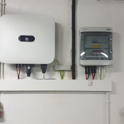 ElektroGriner - Biały falownik Huawei i szara skrzynka elektryczna z bezpiecznikami zamontowane na białej ścianie. Widoczne kable i przewody.