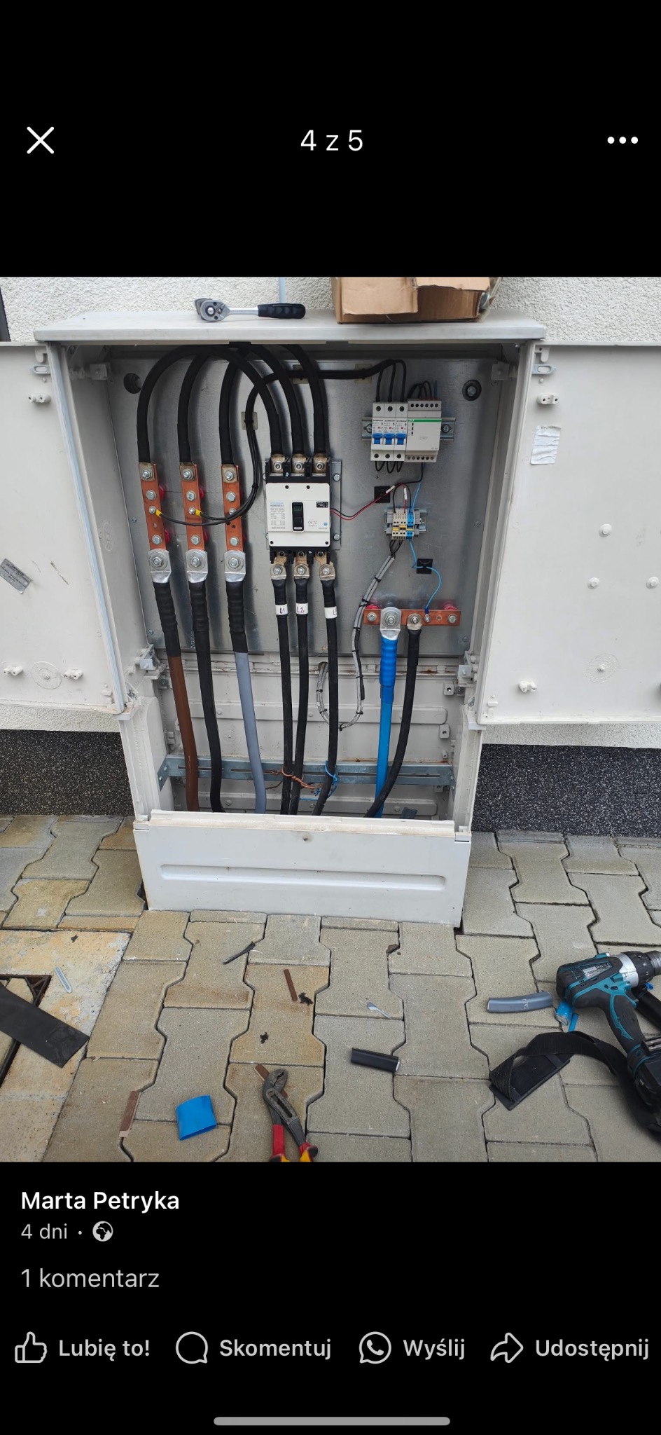 Otwarta skrzynka elektryczna z grubymi kablami, bezpiecznikami i narzędziami na kostce brukowej. Widoczne złącza i elementy instalacji.