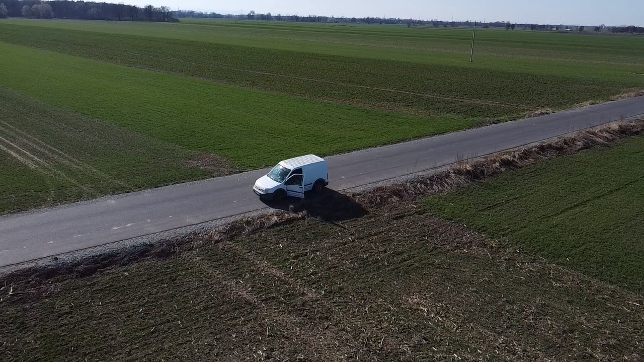 Biały van z otwartymi drzwiami stoi na wąskiej drodze asfaltowej, otoczony polami uprawnymi w słoneczny dzień. Zdjęcie z lotu ptaka.