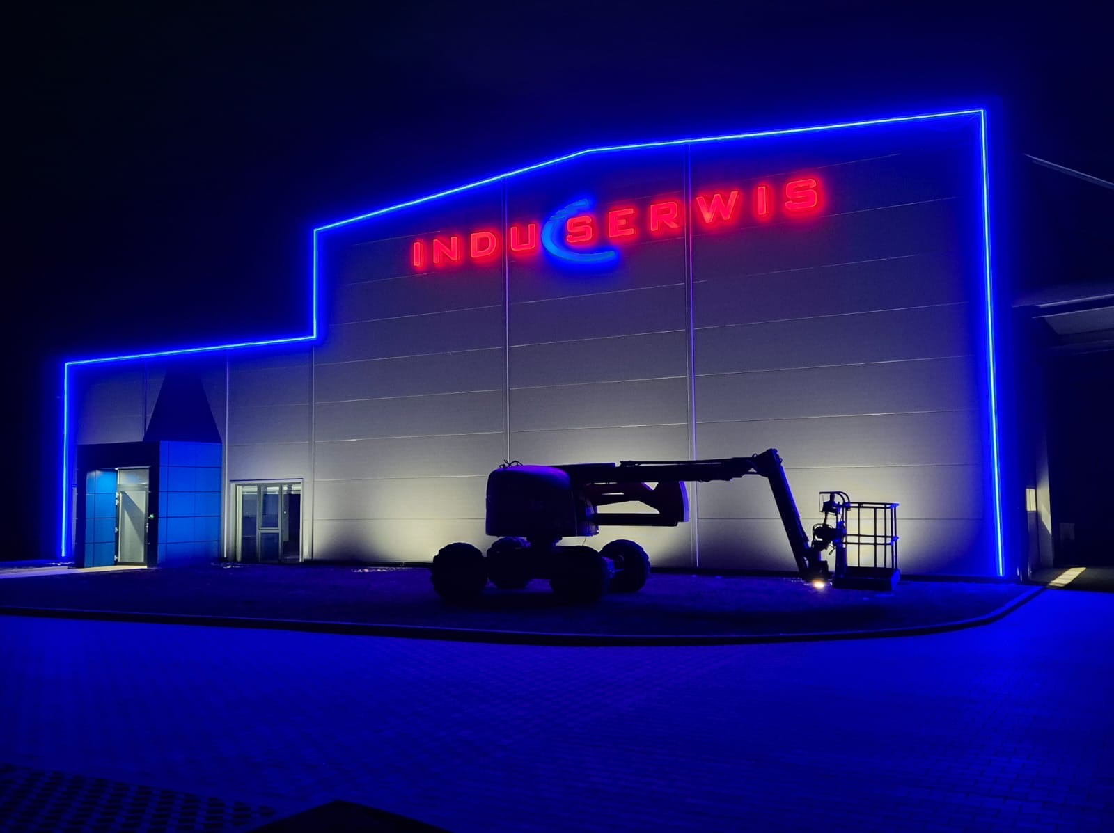 Budynek firmy „Induserwis” z neonowym oświetleniem elewacji i podnośnikiem koszowym na tle nocnego nieba, oświetlony reflektorami.