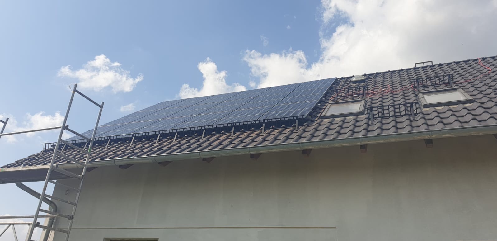 Panele fotowoltaiczne na dachu domu jednorodzinnego, widoczne rusztowania i okna dachowe. Błękitne niebo z chmurami w tle.