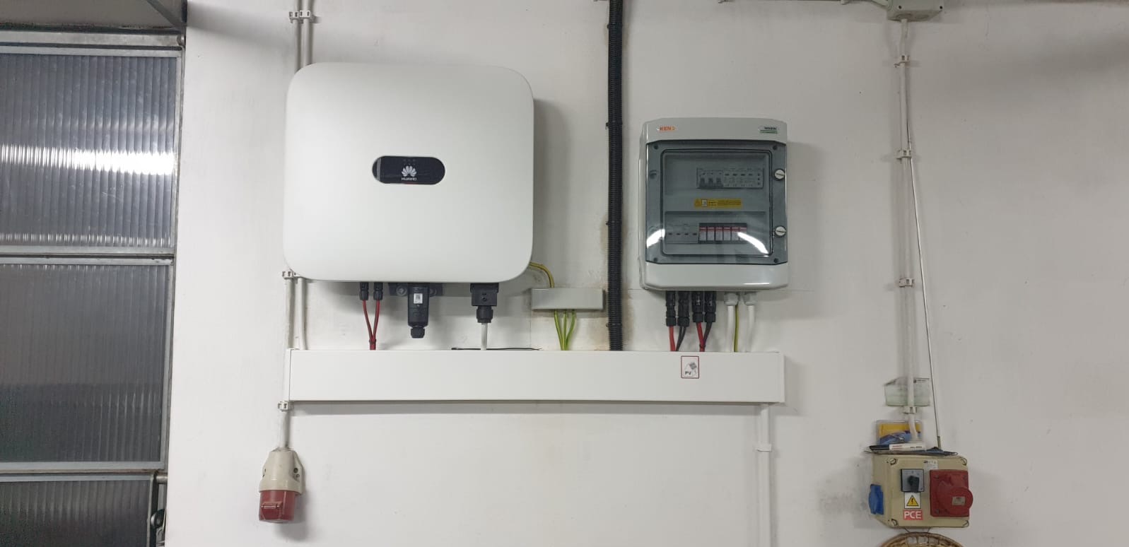 Biały falownik Huawei i szara skrzynka elektryczna z bezpiecznikami zamontowane na białej ścianie. Widoczne kable i przewody.