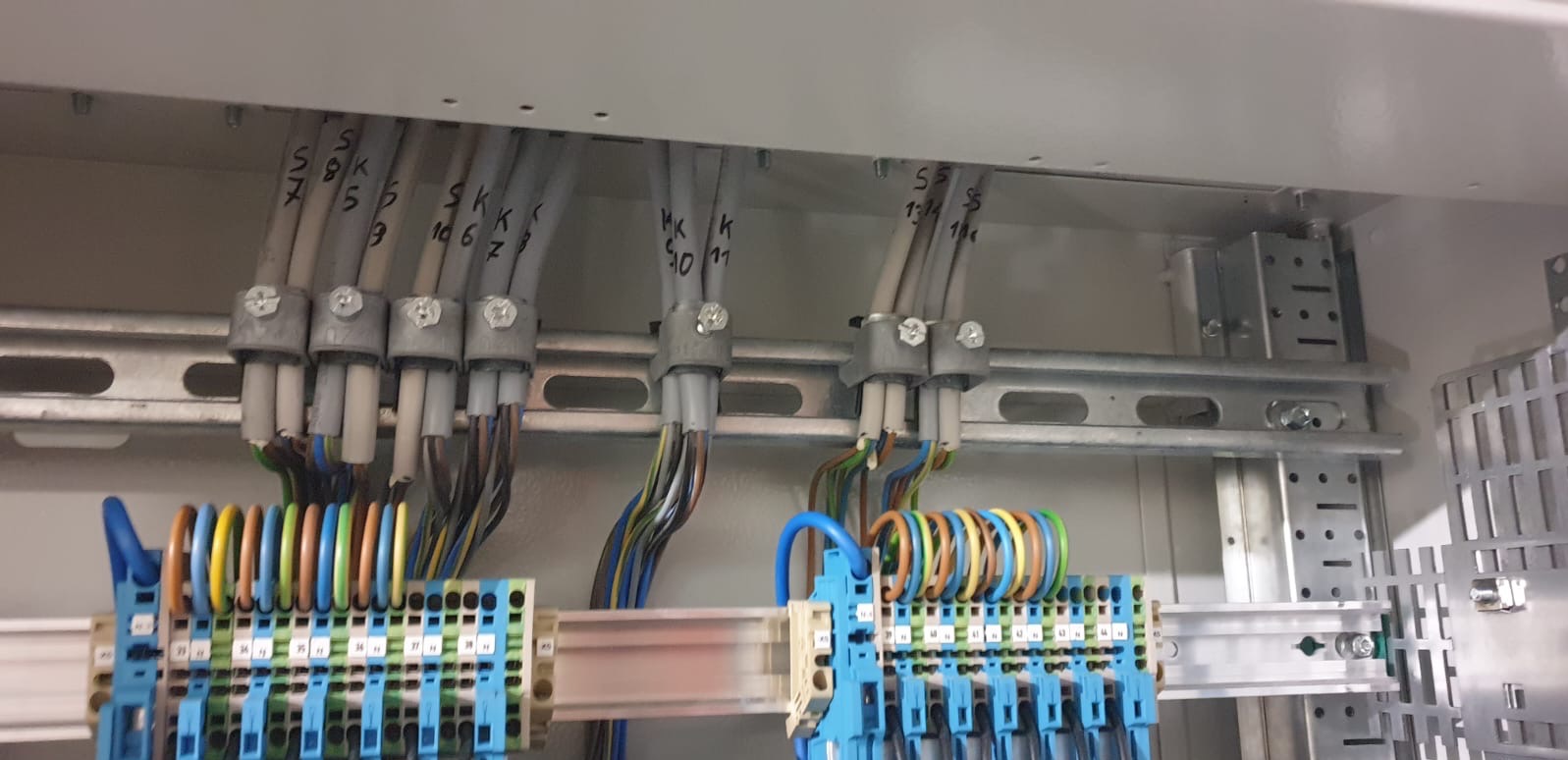 Rozdzielnica elektryczna w trakcie montażu: przewody z oznaczeniami wchodzą do listwy zaciskowej z kolorowymi oznaczeniami. Widoczne metalowe szyny montażowe.