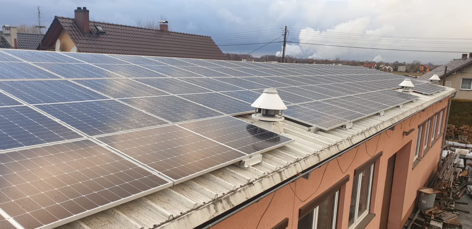 Instalacja fotowoltaiczna na dachu budynku z kominkami wentylacyjnymi, widok z góry, pochmurne niebo w tle.