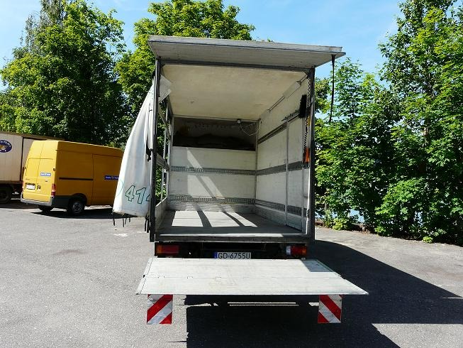 Otwarta pusta przestrzeń ładunkowa samochodu dostawczego z opuszczoną rampą załadunkową, widok z tyłu, na zewnątrz w słoneczny dzień.