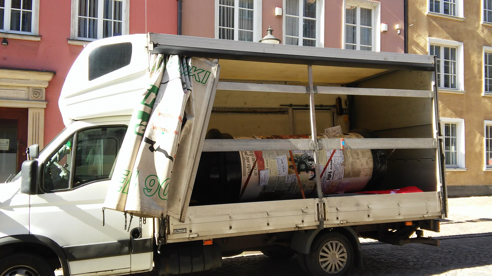 Biały samochód dostawczy z otwartą plandeką, załadowany meblami owiniętymi w gazety, na tle kamienic.