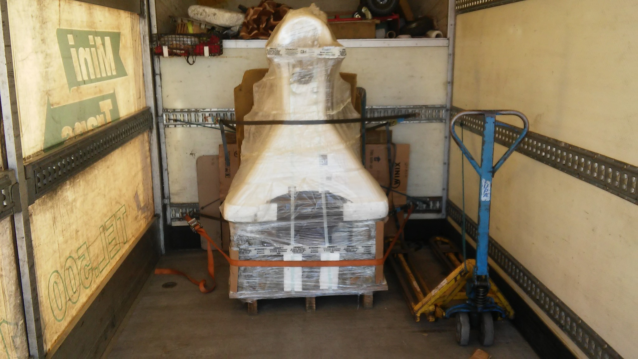 Zabezpieczony, owinięty folią i spięty pasami ładunek, prawdopodobnie kominek, umieszczony na palecie wewnątrz przestrzeni ładunkowej busa, obok wózka paletowego.