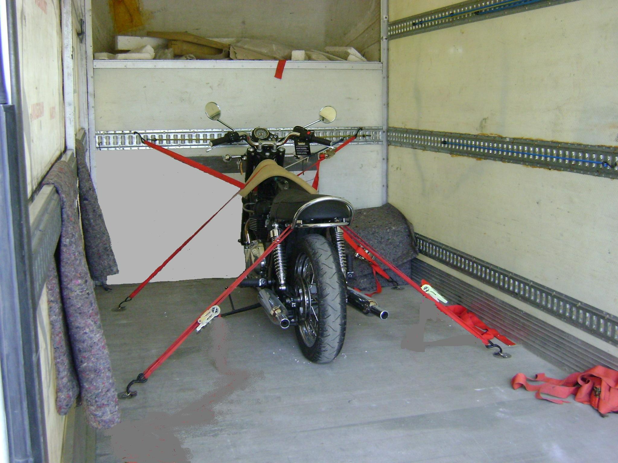 Motocykl zabezpieczony czerwonymi pasami transportowymi wewnątrz przestrzeni ładunkowej busa z metalowymi listwami mocującymi na ścianach.