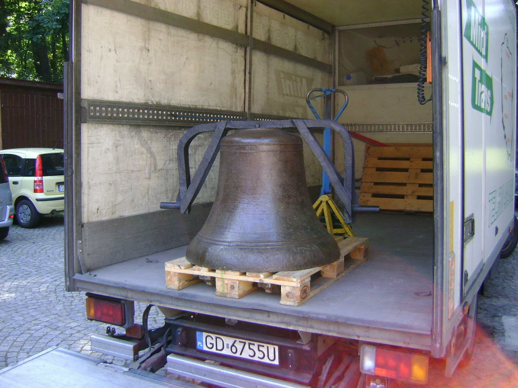Transport dzwonu kościelnego wewnątrz busa dostawczego, zabezpieczonego na palecie drewnianej, widoczny numer rejestracyjny pojazdu.