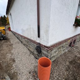 F.H.U. SOWA Joanna Sowa - Przygotowanie terenu wokół domu: warstwa żwiru, rura drenażowa, widoczny fragment fundamentu z ceglaną obudową. W tle sprzęt budowlany i fragment elewacji.