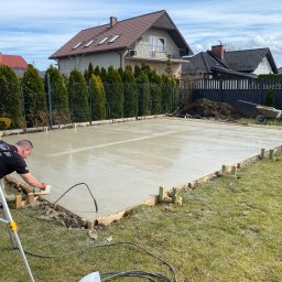 F.H.U. SOWA Joanna Sowa - Pracownik w czarnej koszulce wygładza świeżo wylaną betonową płytę na posesji, widoczny laserowy niwelator na statywie, drewniane szalunki i domy w tle.