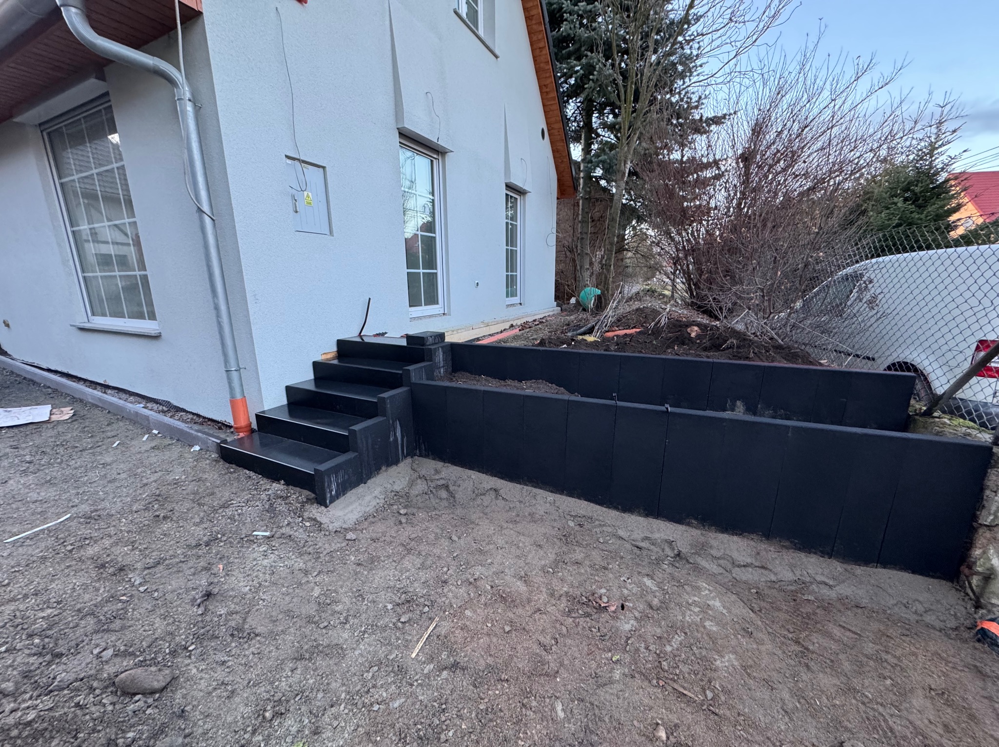 Nowoczesne schody wejściowe z czarnego kamienia prowadzące do domu z podwyższonym tarasem, otoczone ziemią i roślinnością. Minimalistyczny design.