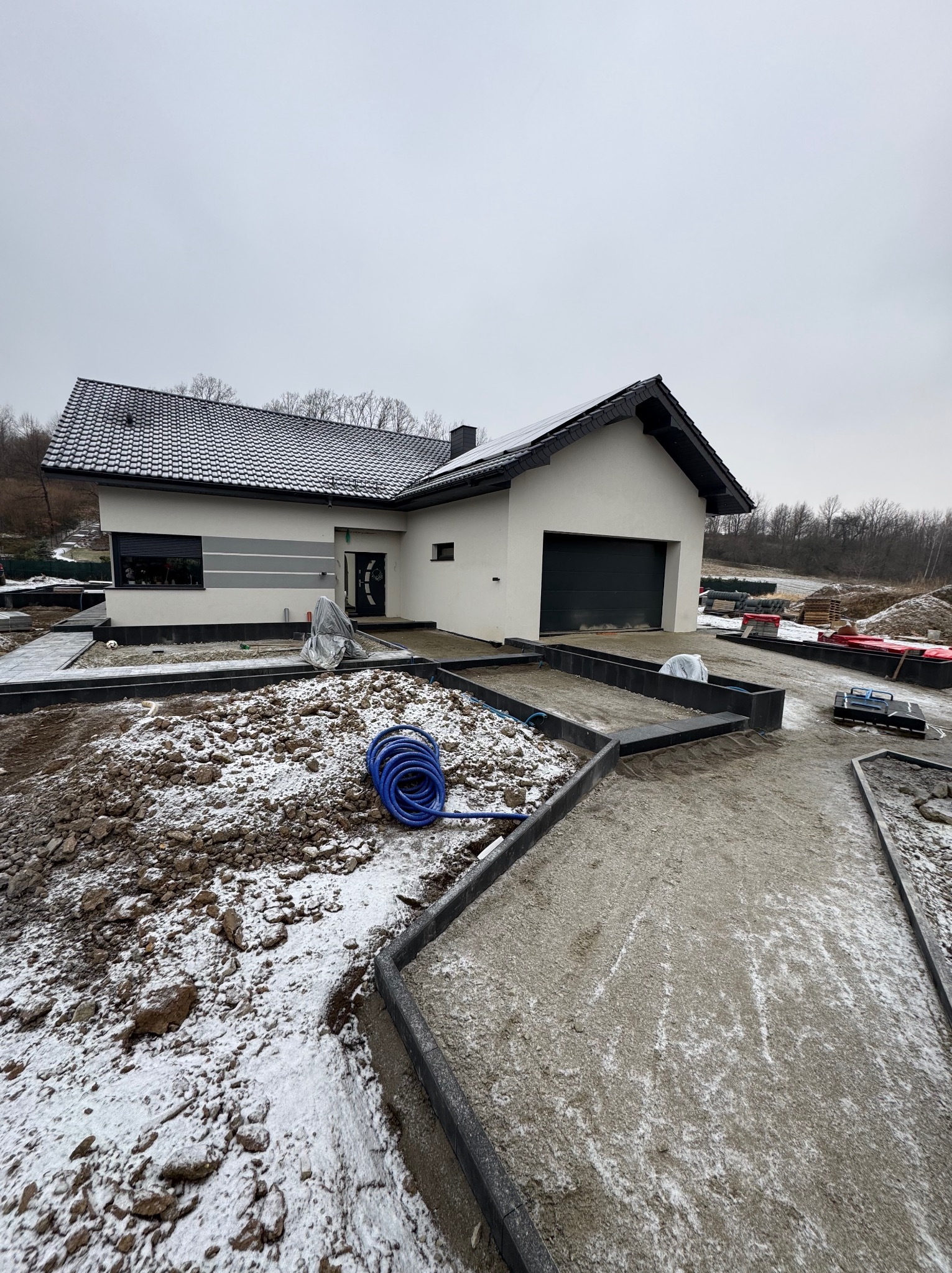 Nowoczesny dom jednorodzinny z przygotowanym podłożem pod kostkę brukową i obrzeżami. Widoczny zwój niebieskiego węża ogrodowego. Pochmurny dzień.