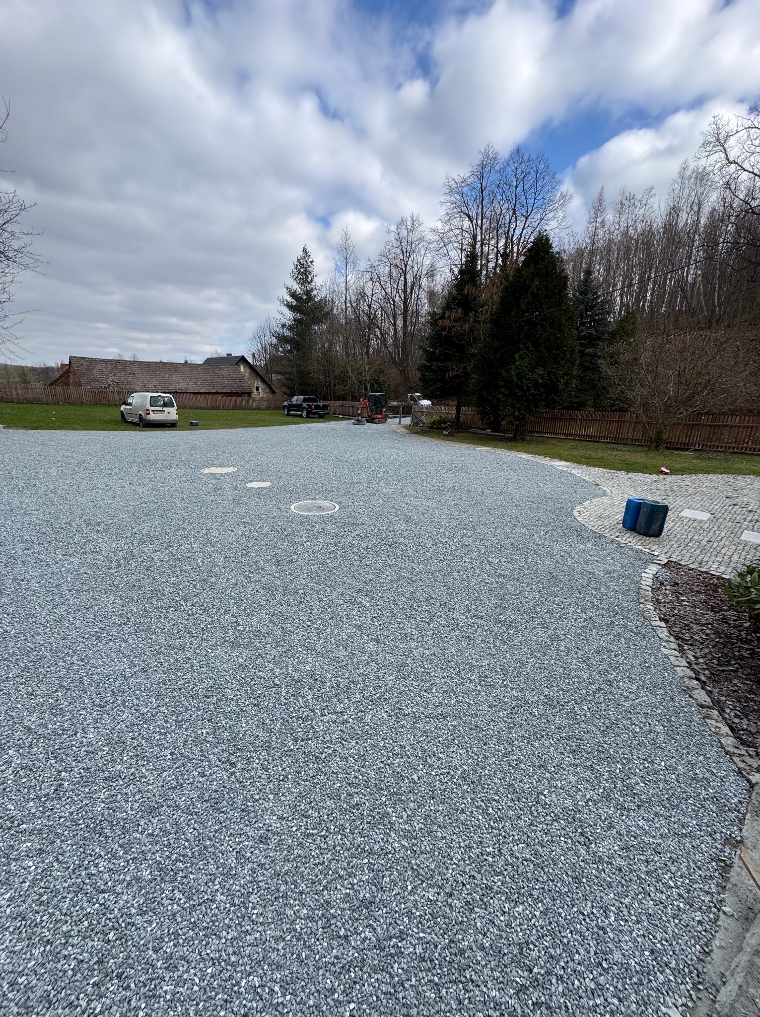 Utwardzona droga z szarego kruszywa na posesji. W tle budynek, drzewa i błękitne niebo z chmurami. Widoczne studzienki kanalizacyjne i zaparkowane pojazdy.