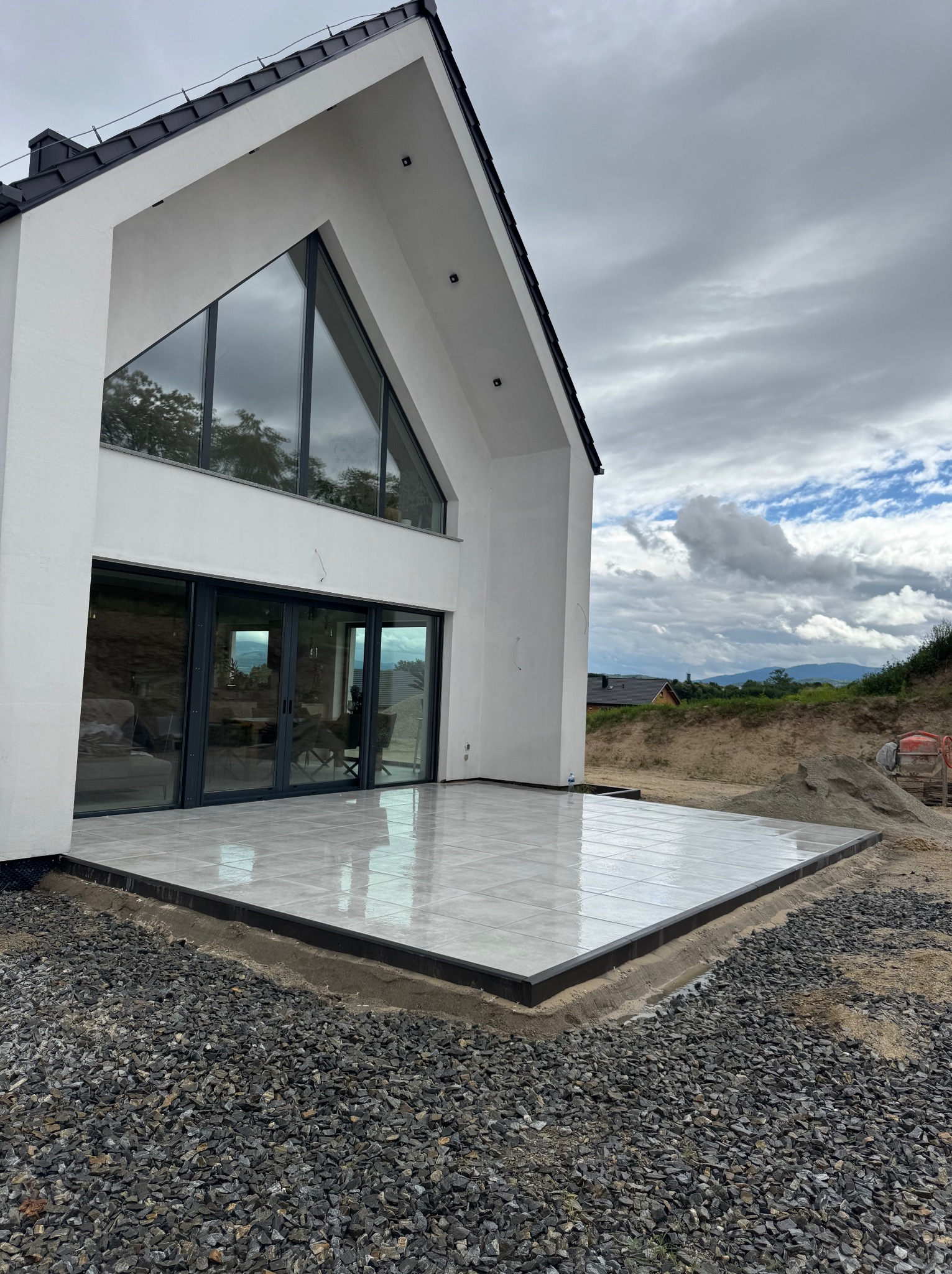 Nowoczesny taras z jasnych płytek, obrzeża z ciemnego kamienia, na tle białego domu z dużymi przeszkleniami i szarym niebem. Widoczne przygotowania do dalszych prac.