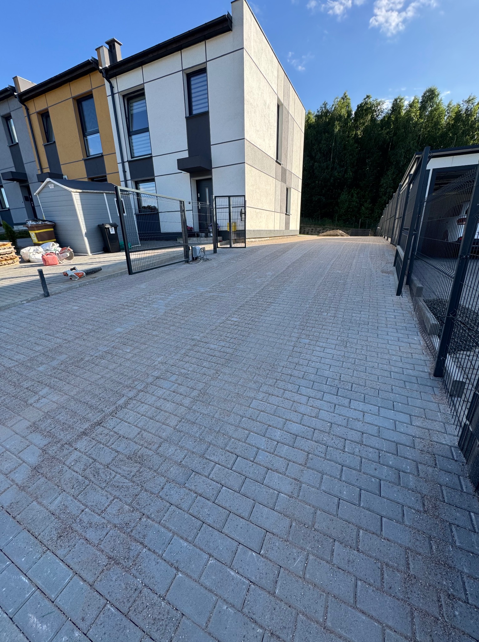 Ułożona, szara kostka brukowa na podjeździe w Ożarach, z widocznym ogrodzeniem panelowym i fragmentem nowoczesnego domu w tle. Widok z perspektywy.