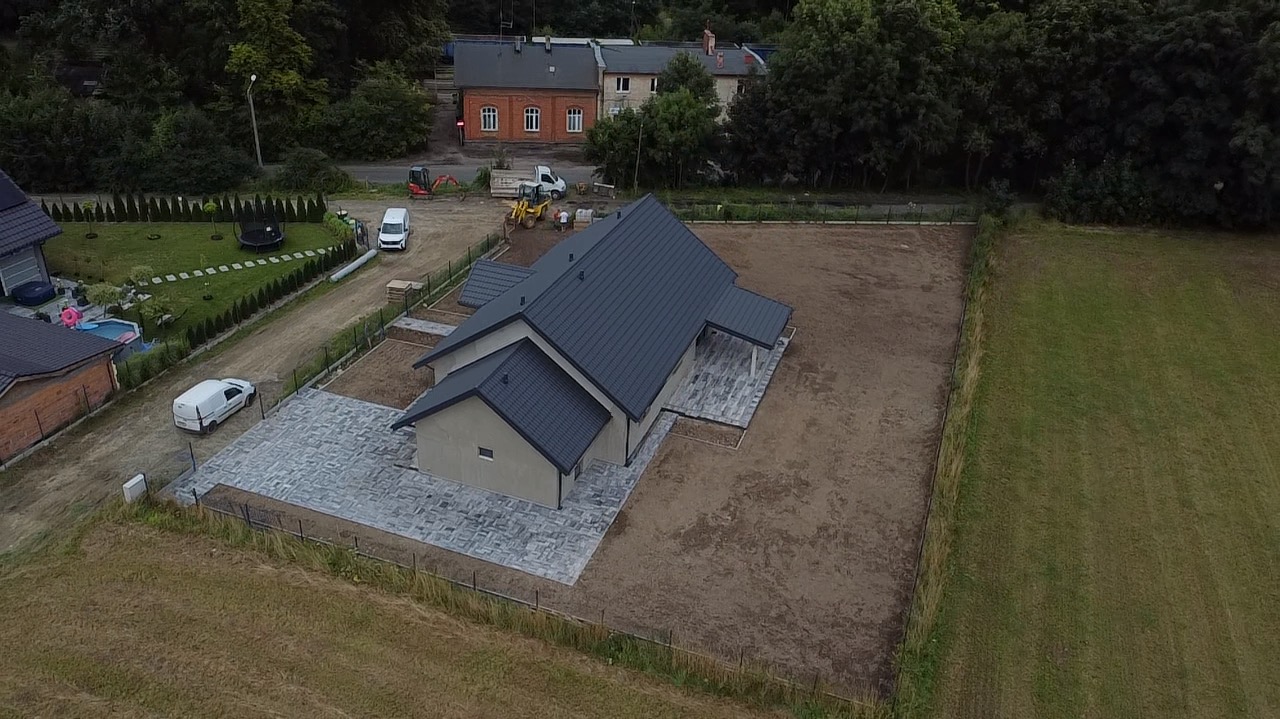 Nowoczesny dom z ciemnym dachem i szarą kostką brukową na podjeździe, widok z drona. Częściowo zagospodarowany teren, w tle budynki i zieleń.
