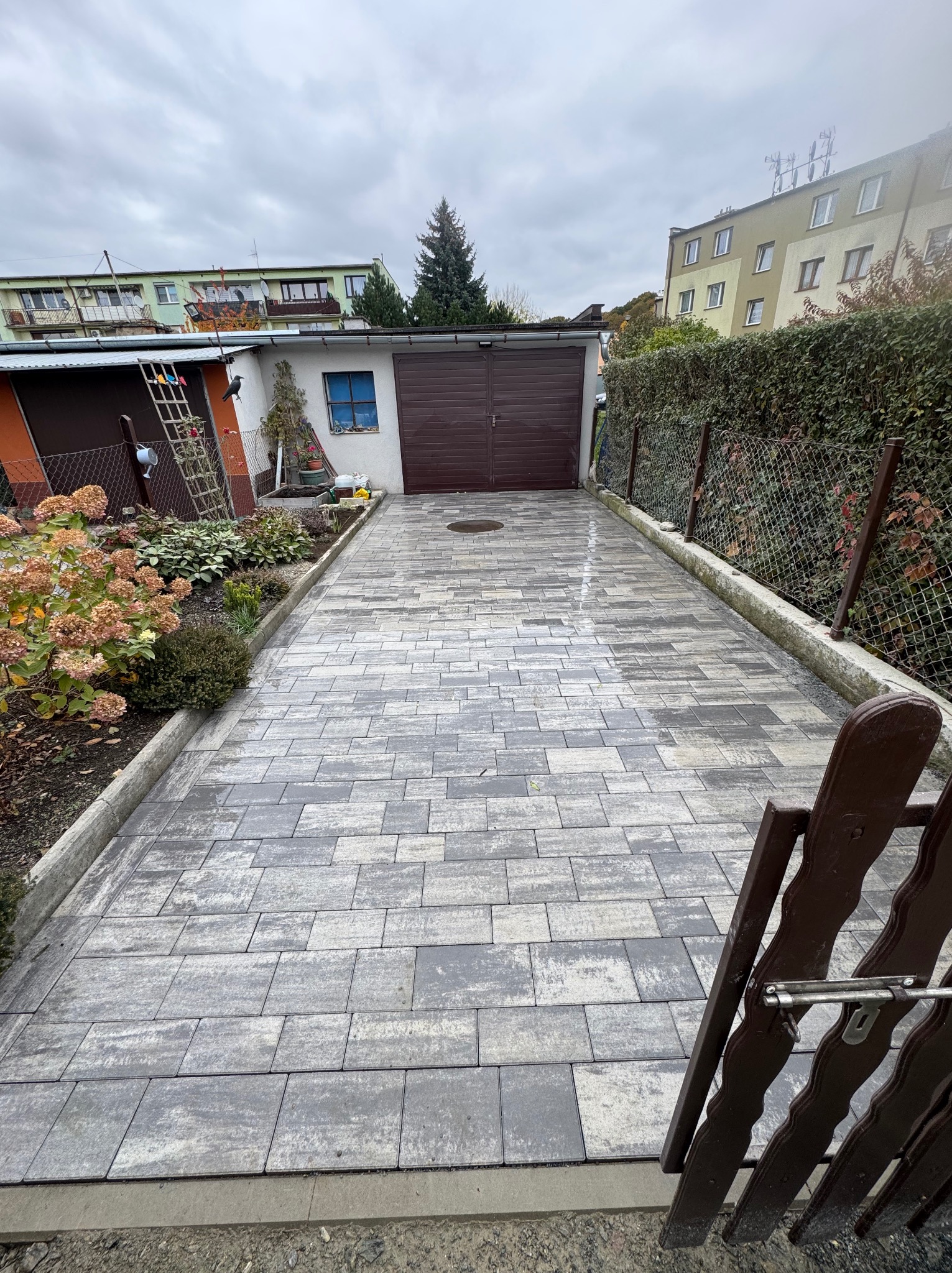 Ułożona szara kostka brukowa na podjeździe do garażu, widok z perspektywy furtki. W tle budynki mieszkalne i zieleń. Kostka ułożona w regularny wzór.