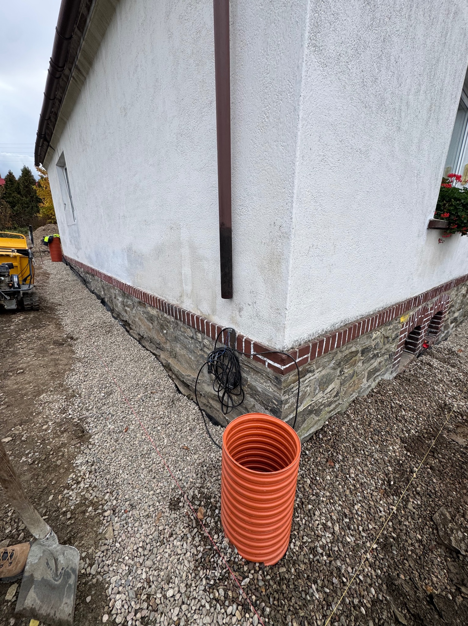 Przygotowanie terenu wokół domu: warstwa żwiru, rura drenażowa, widoczny fragment fundamentu z ceglaną obudową. W tle sprzęt budowlany i fragment elewacji.