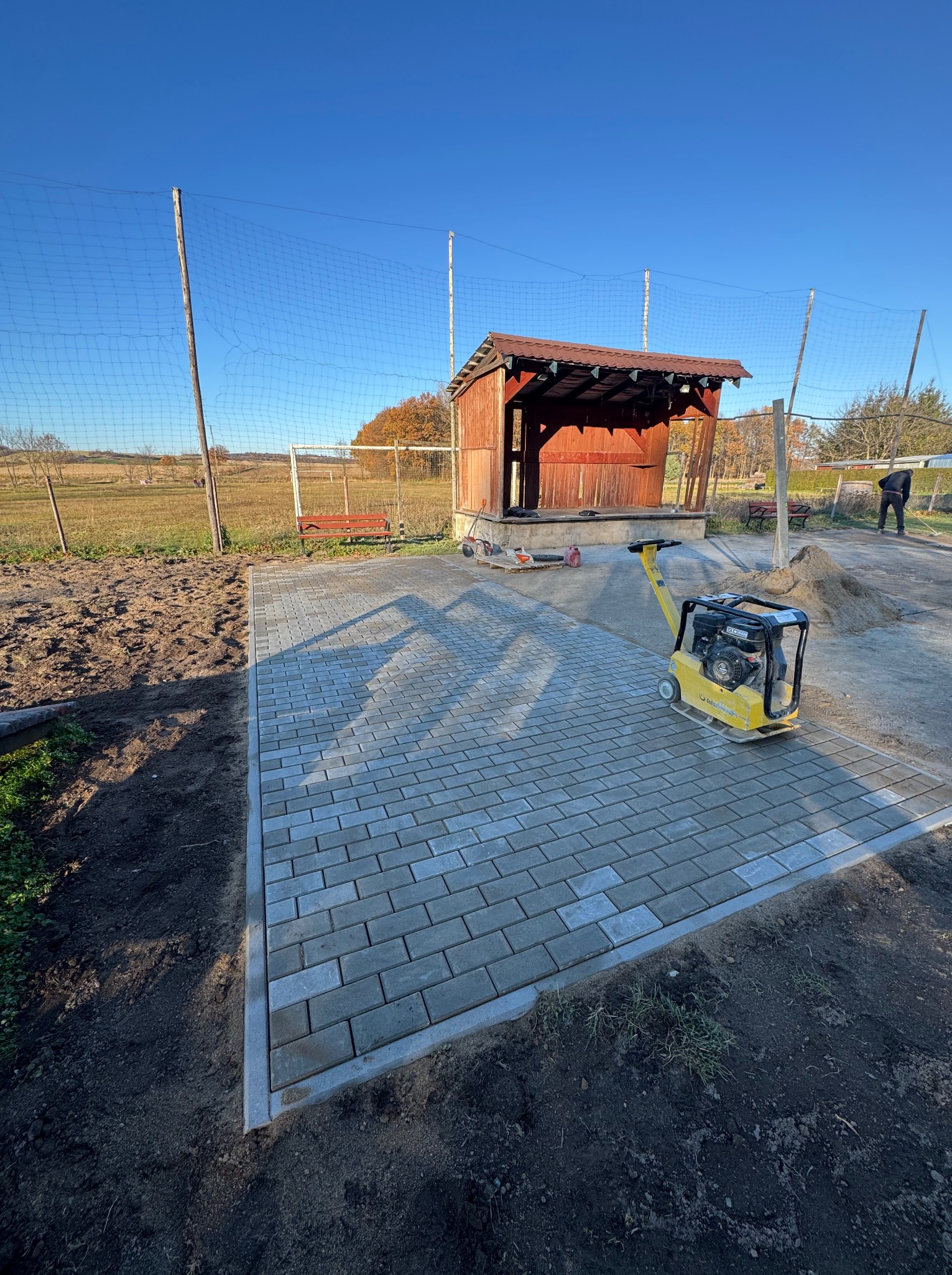 Szara kostka brukowa ułożona na placu, żółta zagęszczarka obok, w tle drewniana wiata i siatka ogrodzeniowa na tle błękitnego nieba. Częściowo wykończona nawierzchnia.