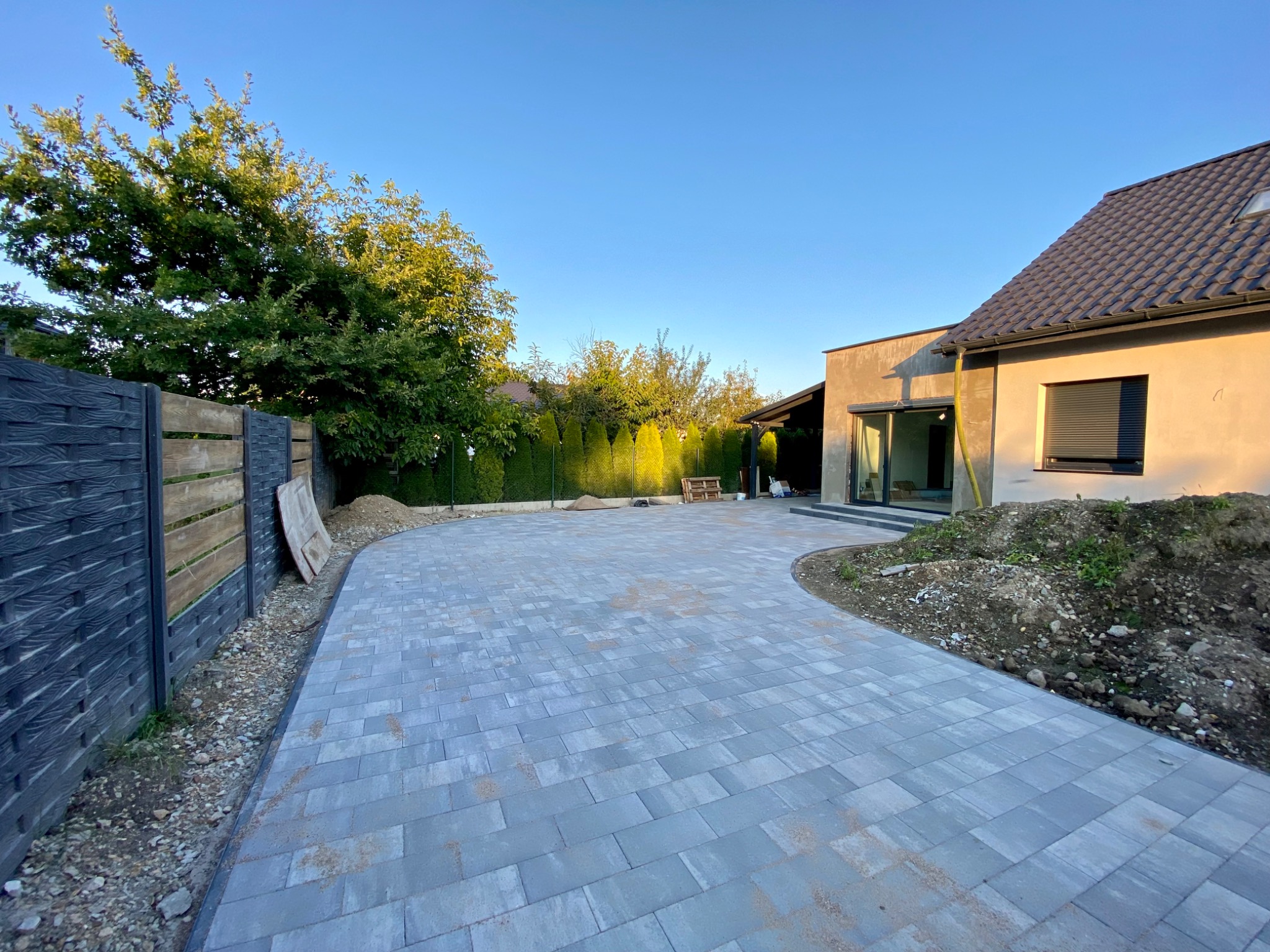 Nowo ułożona, szara kostka brukowa na podjeździe przed nowoczesnym domem z ciemną stolarką okienną, widoczne drzewa i czarno-drewniane ogrodzenie.