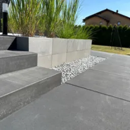 Nowoczesne schody zewnętrzne z ciemnych, betonowych stopni prowadzące na taras z szarych płyt, z podwyższoną rabatą z jasnych betonowych bloczków, wypełnioną ozdobną trawą i białym grysem.