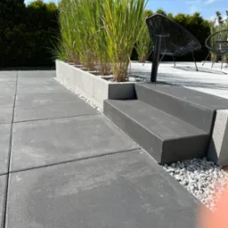 Nowoczesny taras z szarych płyt, dwustopniowe schody z ciemnego betonu, donice z trawą ozdobną i czarne krzesła w tle.